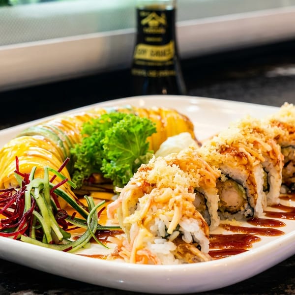 Two sushi rolls, one topped with crispy flakes, on a white plate with soy sauce in the background.