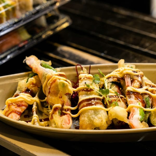 Tempura sushi rolls drizzled with mayonnaise and dark sauce, garnished with microgreens on a boat-shaped plate.